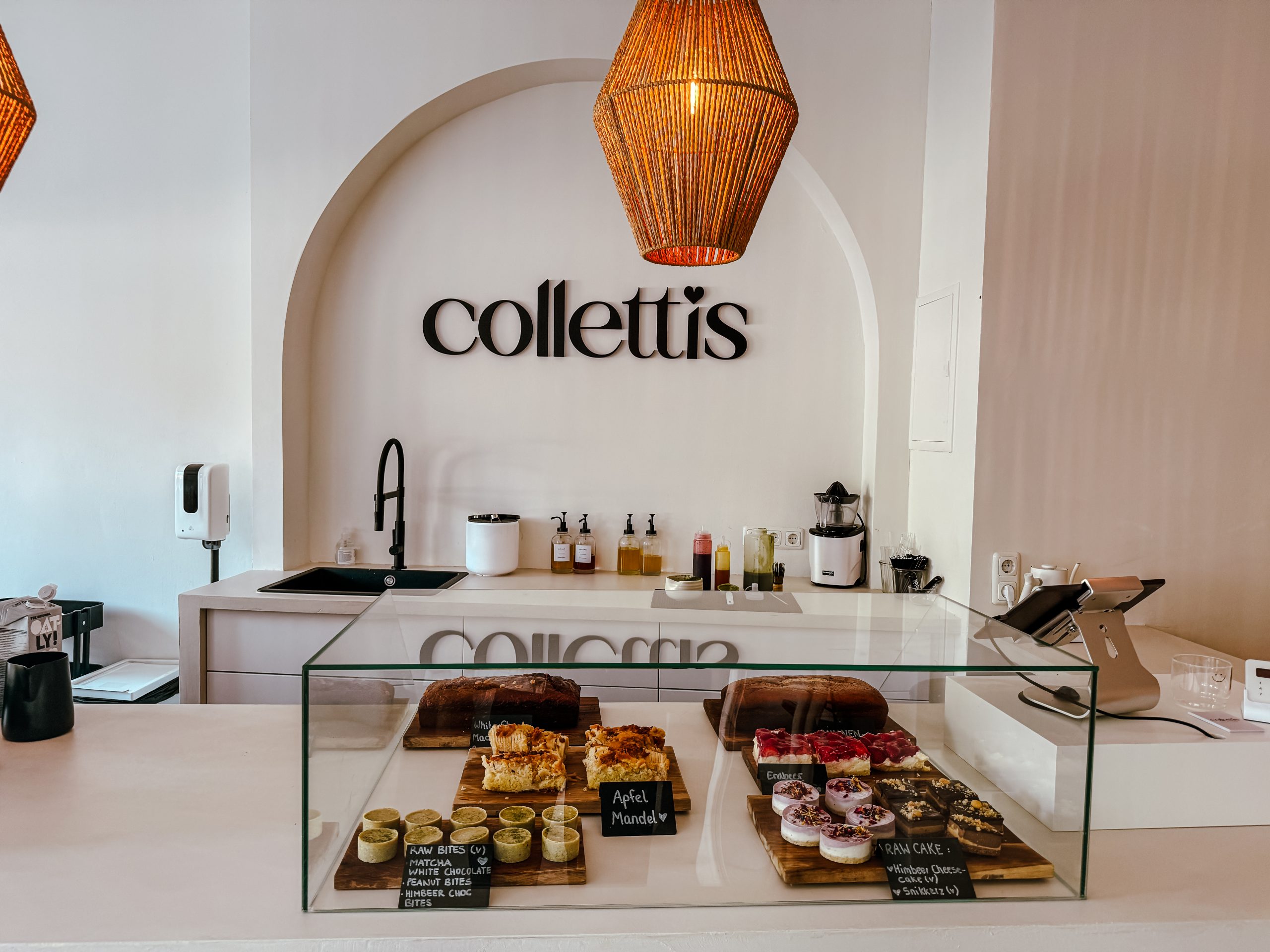 Eine moderne Bäckertheke mit Backwaren in einer Glasvitrine, darüber Pendelleuchten aus Korbgeflecht und dem Schriftzug Colletti's an der Wand hinter der Theke.