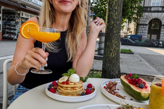 Eine lächelnde Frau sitzt an einem Cafétisch im Freien mit einem Stapel Pfannkuchen, Obst und Avocado-Toast und hält einen Orangencocktail in der Hand. Im Hintergrund sind Gebäude, Bäume und andere Speisen zu sehen.