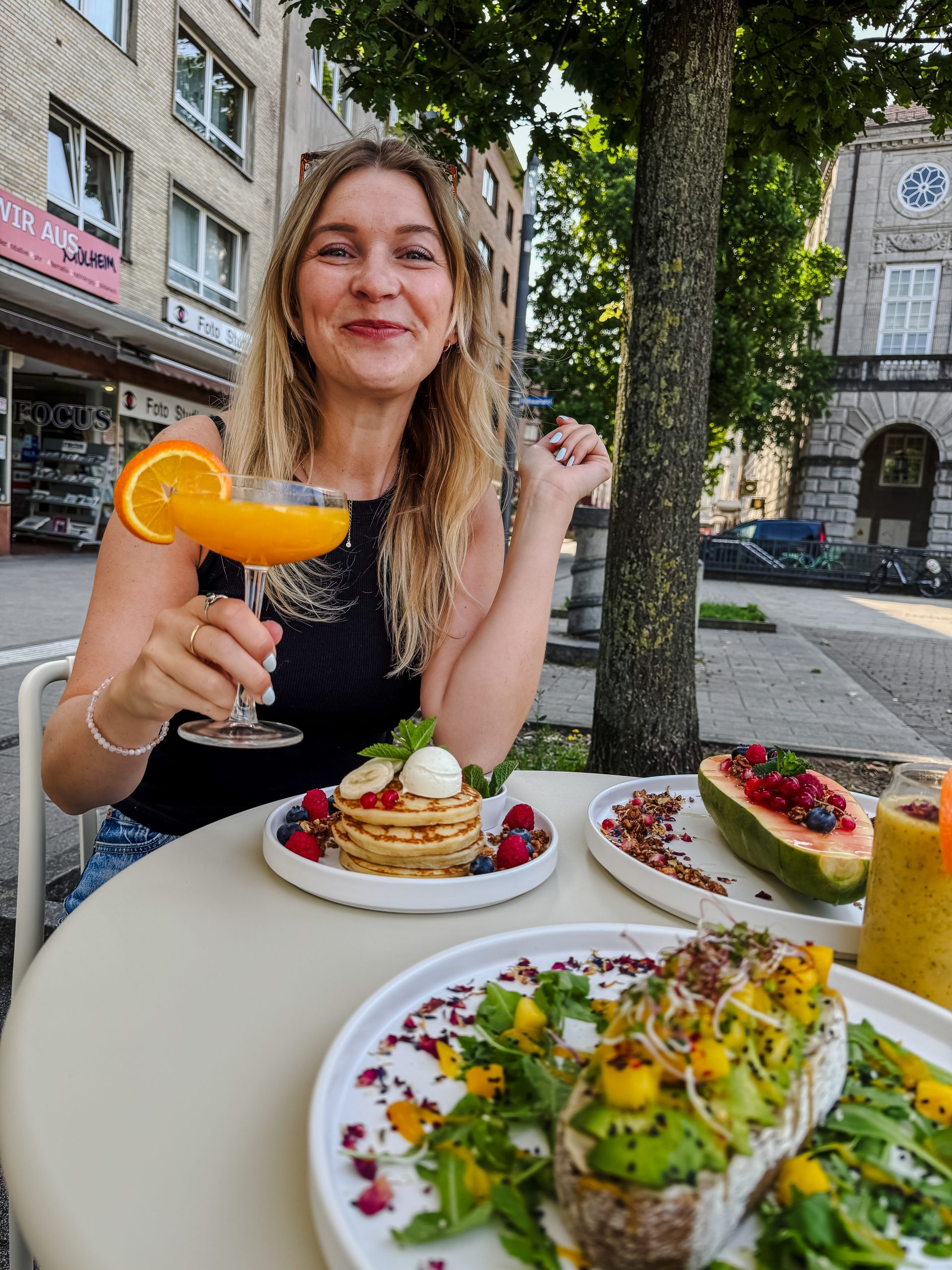 Eine lächelnde Frau sitzt an einem Cafétisch im Freien mit einem Stapel Pfannkuchen, Obst und Avocado-Toast und hält einen Orangencocktail in der Hand. Im Hintergrund sind Gebäude, Bäume und andere Speisen zu sehen.