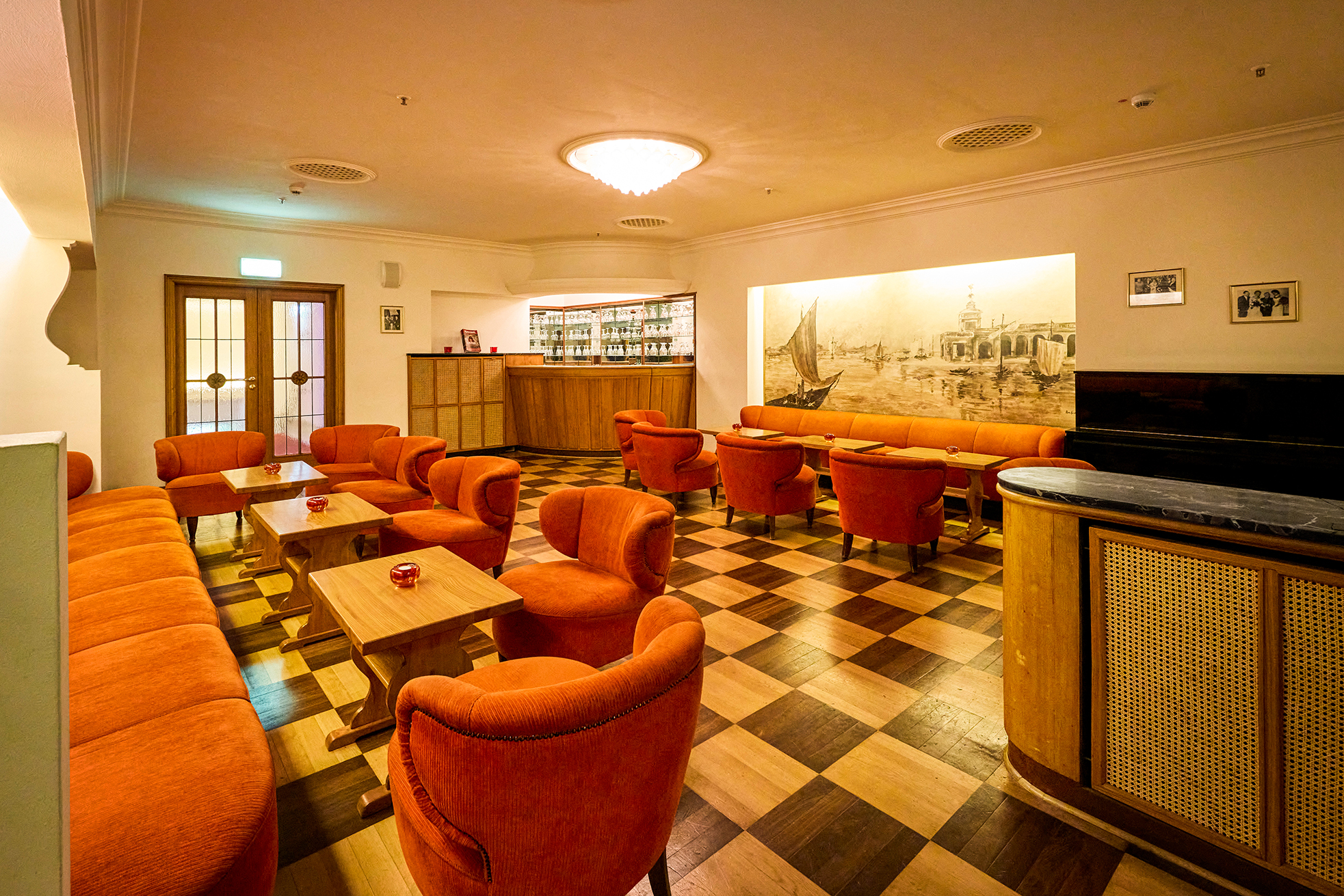 Eine gemütliche Lounge mit orangefarbenen Sesseln und Holztischen auf einem karierten Boden. Im hinteren Teil befindet sich eine Bar mit Flaschenregalen, die Wand ist mit einem Wandbild geschmückt. Die warme Beleuchtung schafft eine einladende Atmosphäre.