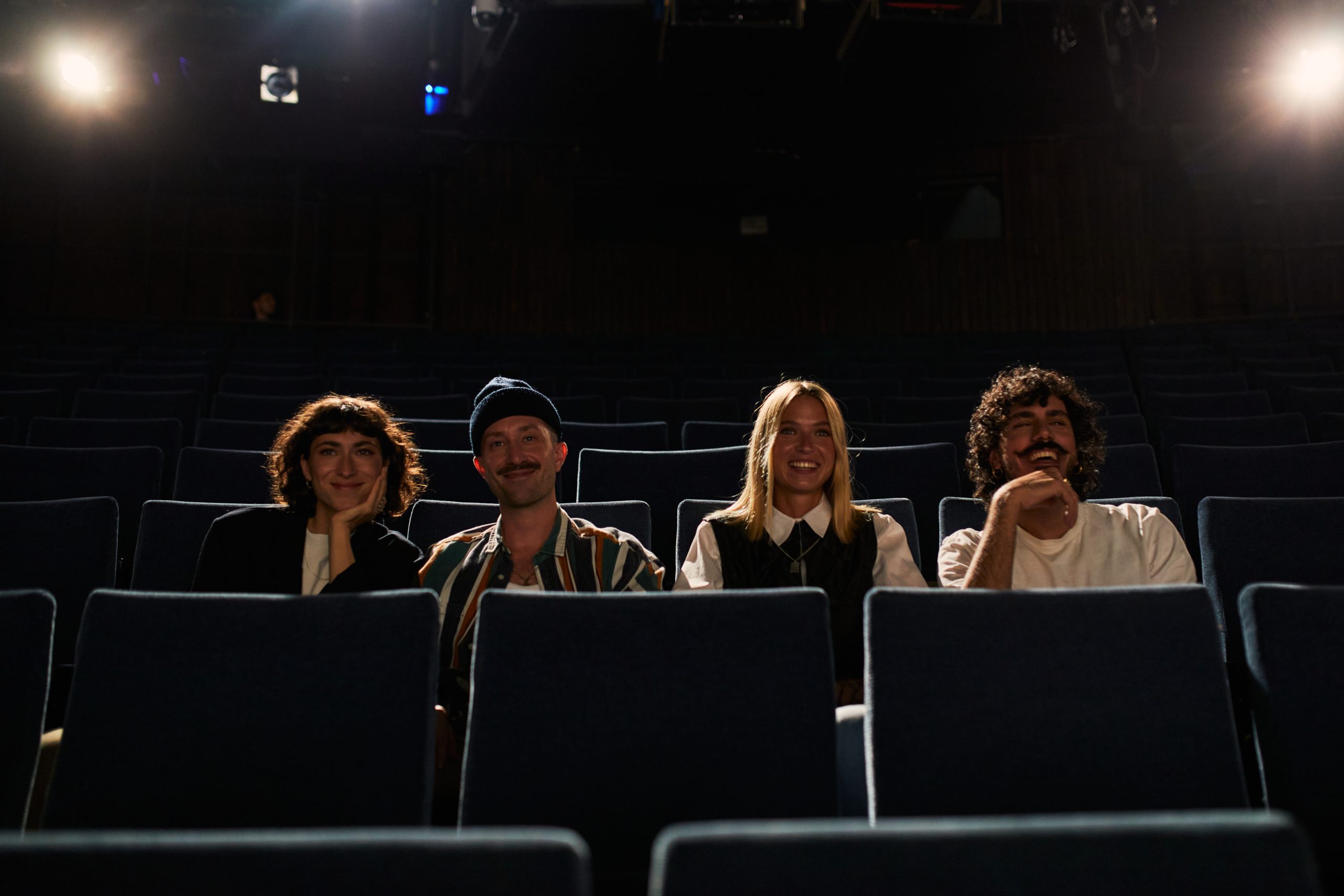Vier Personen sitzen in der ersten Reihe eines dunklen, leeren Theaters, lächeln und schauen in die Kamera. Zwei Männer und zwei Frauen sitzen gleichmäßig auf den Sitzen verteilt und werden von sanften Bühnenscheinwerfern beleuchtet.