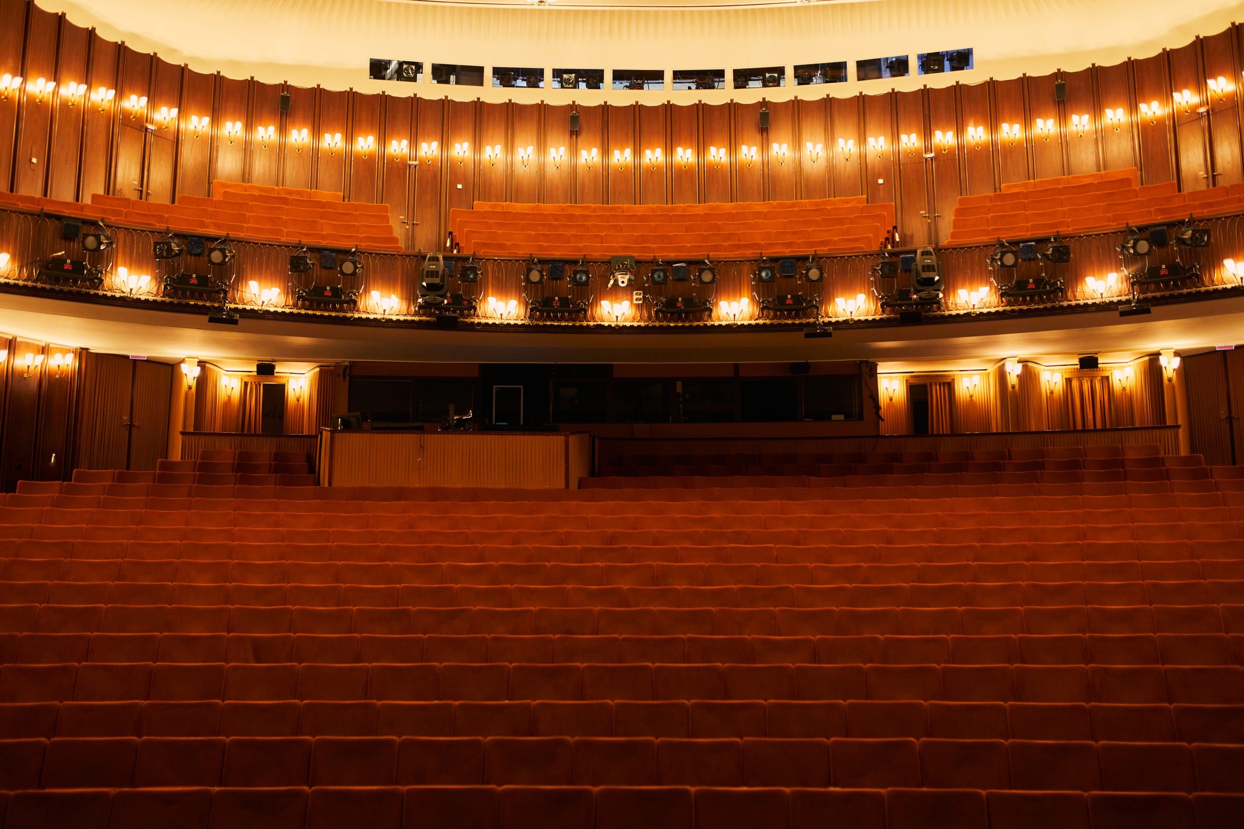 Ein leerer Theatersaal mit Sitzreihen aus rotem Samt, Holzvertäfelung, leuchtenden Lichtern auf den Balkonen und einer nicht einsehbaren Bühne. Die Atmosphäre ist warm und einladend, aber ruhig.