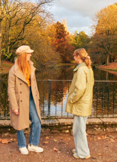 Zwei Menschen in Herbstmänteln stehen an einem schwarzen Metallzaun mit Blick auf einen Teich, umgeben von bunten Herbstbäumen mit orangefarbenen und gelben Blättern, die den Boden bedecken.