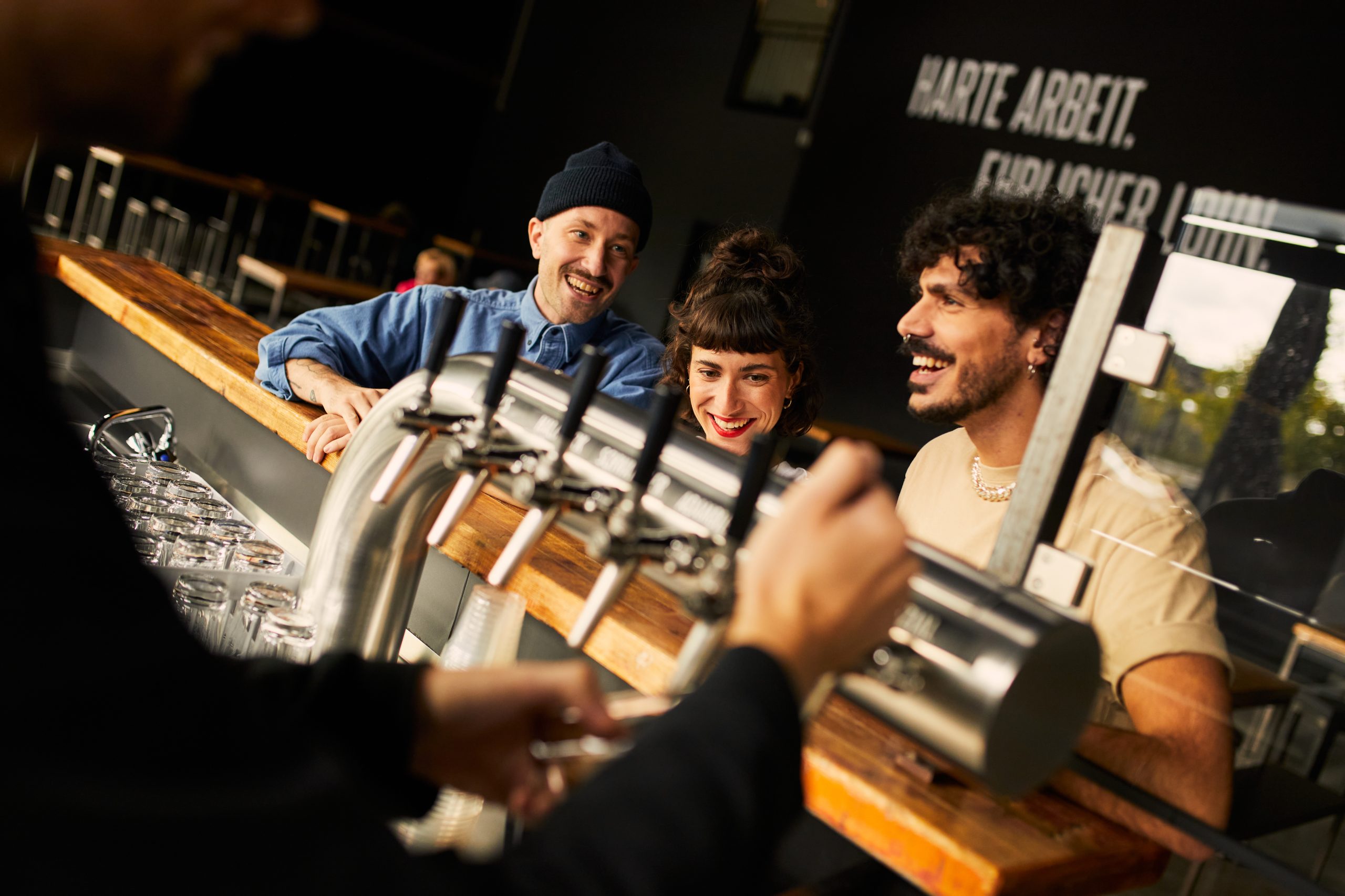 Drei Personen an einer Bar lächeln und unterhalten sich, während ein Barkeeper Getränke aus einer Reihe von Zapfhähnen einschenkt. Die Atmosphäre ist lebhaft und freundlich, die Gläser stehen aufgereiht auf dem Tresen.