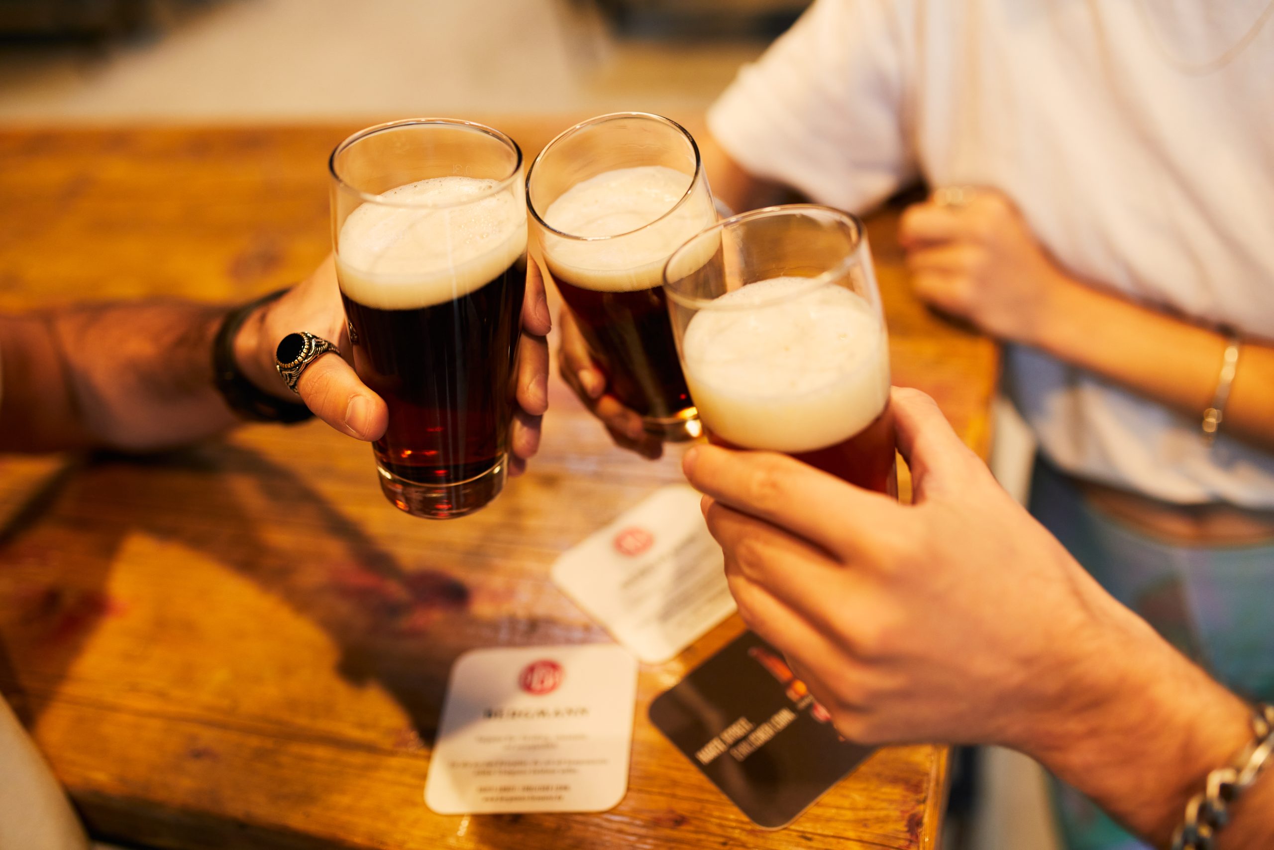 Drei Personen stoßen an einem Holztisch mit Gläsern mit dunklem Bier an, jeder hält ein Glas mit schaumiger Schaumkrone; im Hintergrund sind Getränkeuntersetzer und verschwommene Arme zu sehen.