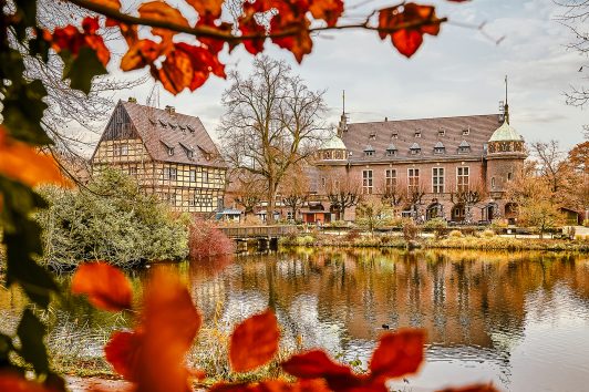 Ein malerischer Blick auf historische Gebäude an einem ruhigen Teich, umrahmt von Herbstlaub. Unter einem bewölkten Himmel spiegeln sich die Gebäude und Bäume im Wasser.