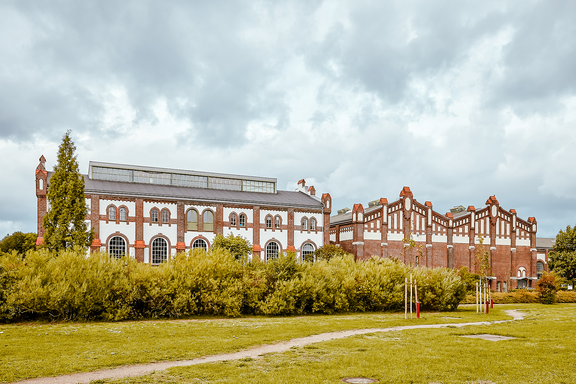 Ein großes, historisches Backsteingebäude mit gewölbten Fenstern und dekorativen Mustern liegt hinter grünen Büschen und einem grasbewachsenen Feld unter einem bewölkten Himmel.