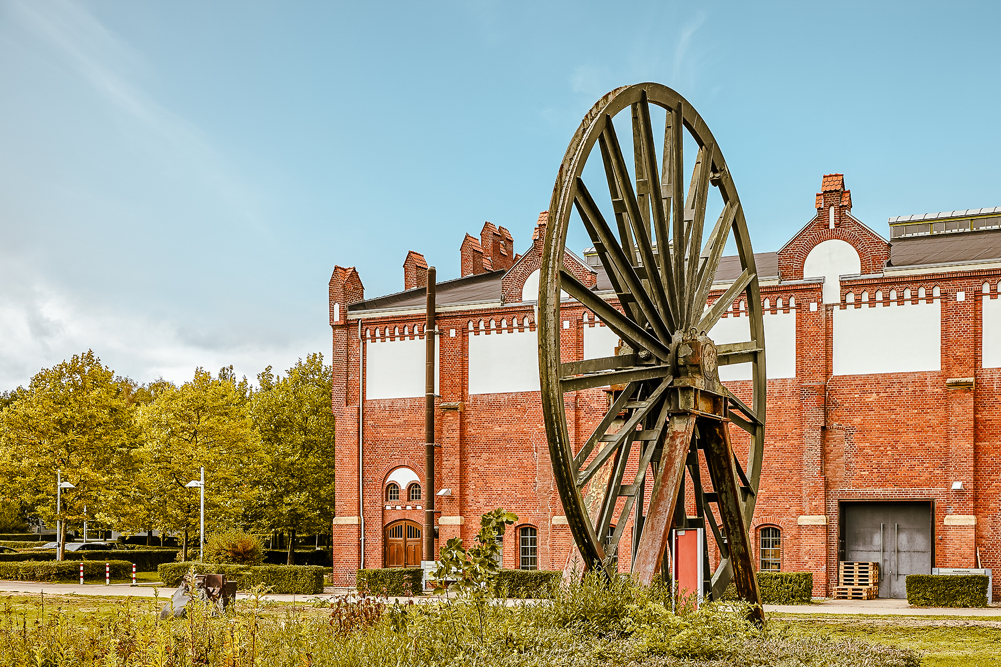 Ein großes, altes Holzrad steht aufrecht vor einem historischen Industriegebäude aus rotem Backstein, umgeben von Grünflächen und Bäumen unter einem klaren blauen Himmel.