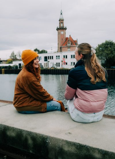 Zwei Frauen sitzen auf einem Betonvorsprung am Wasser, schauen sich an und lächeln. Die eine trägt eine gelbe Mütze und einen braunen Mantel, die andere eine rosa und marineblaue Jacke. Im Hintergrund sind ein Uhrenturm und Gebäude zu sehen.