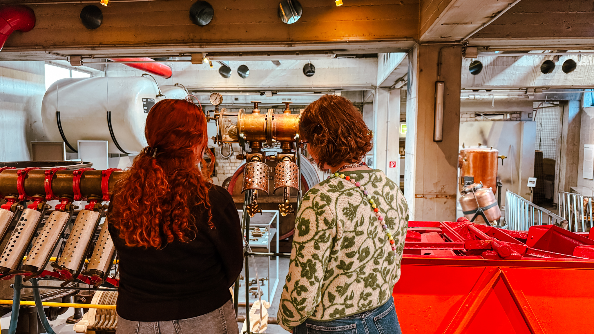 Zwei Personen mit lockigem Haar, von hinten gesehen, stehen in einem Industriemuseum und betrachten alte Maschinen und Metallgeräte in einem hell erleuchteten, geräumigen Raum.