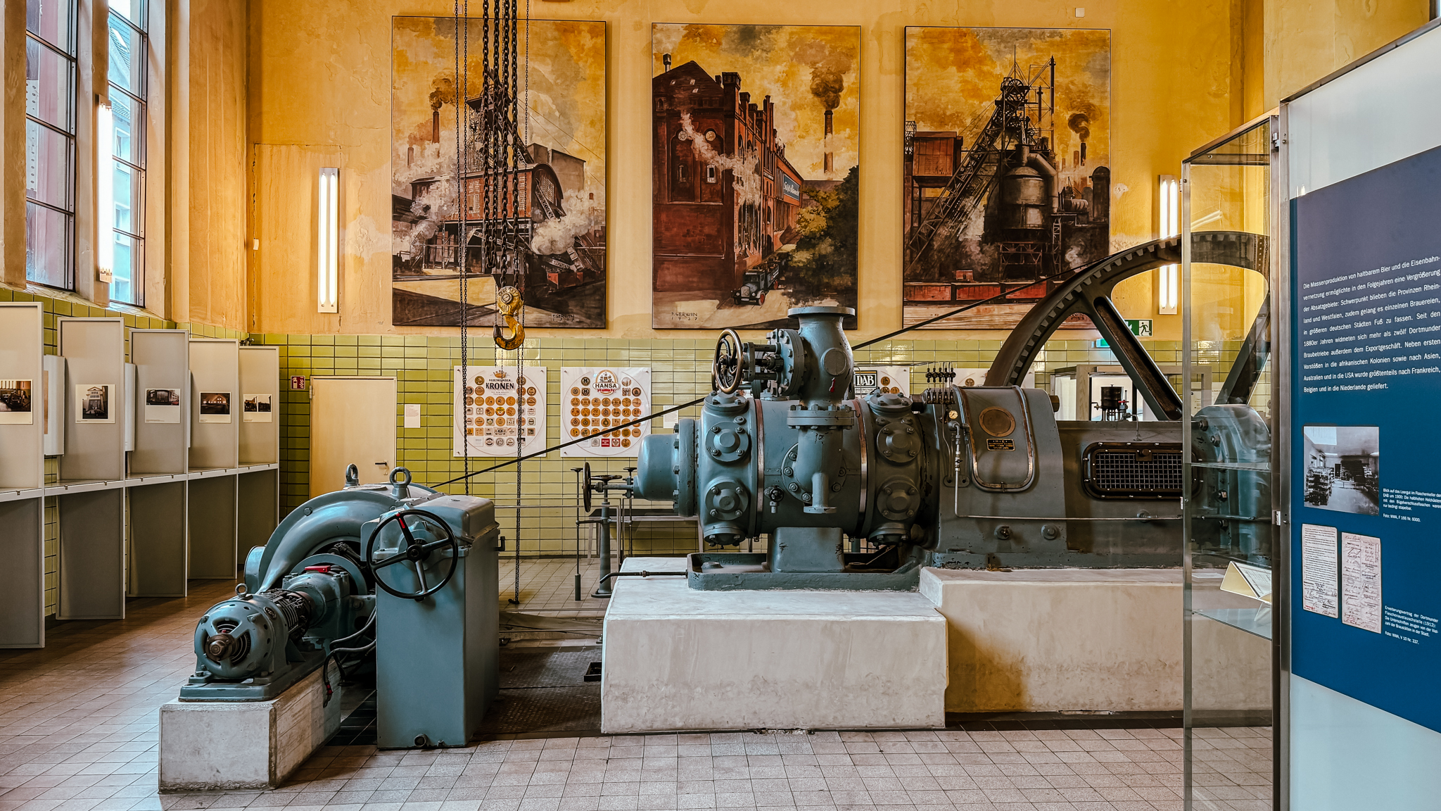 In einem Museumsraum sind große alte Industriemaschinen ausgestellt. An den gelben Wänden hängen historische Fotos und Kunstwerke zum Thema Industrie. Gläserne Exponate und Informationstafeln sind in dem Raum verteilt.