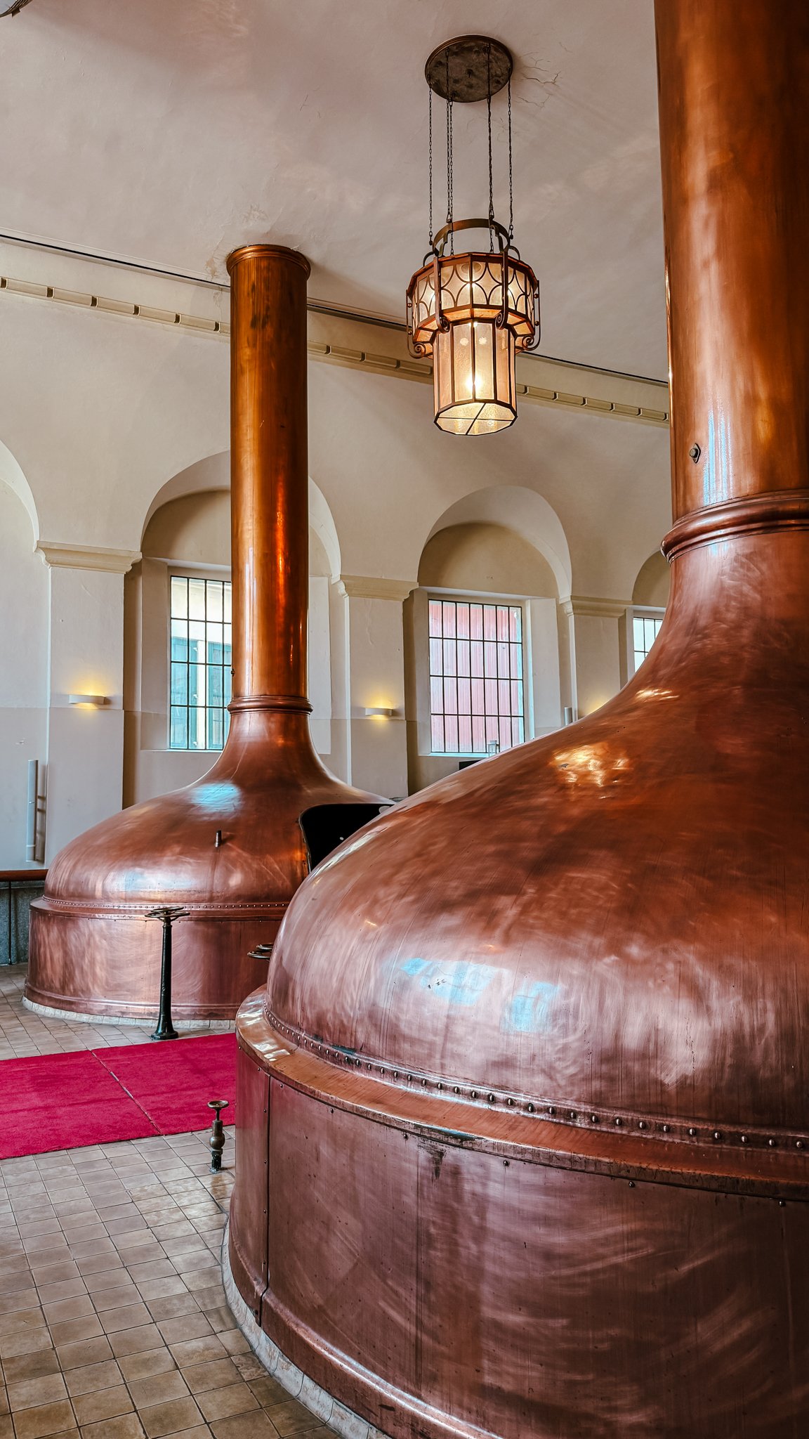 Große kupferne Braukessel stehen unter Bogenfenstern und einer hängenden Laterne in einem geräumigen, hellen Brauereiraum mit gefliesten Böden und einem roten Teppichläufer.