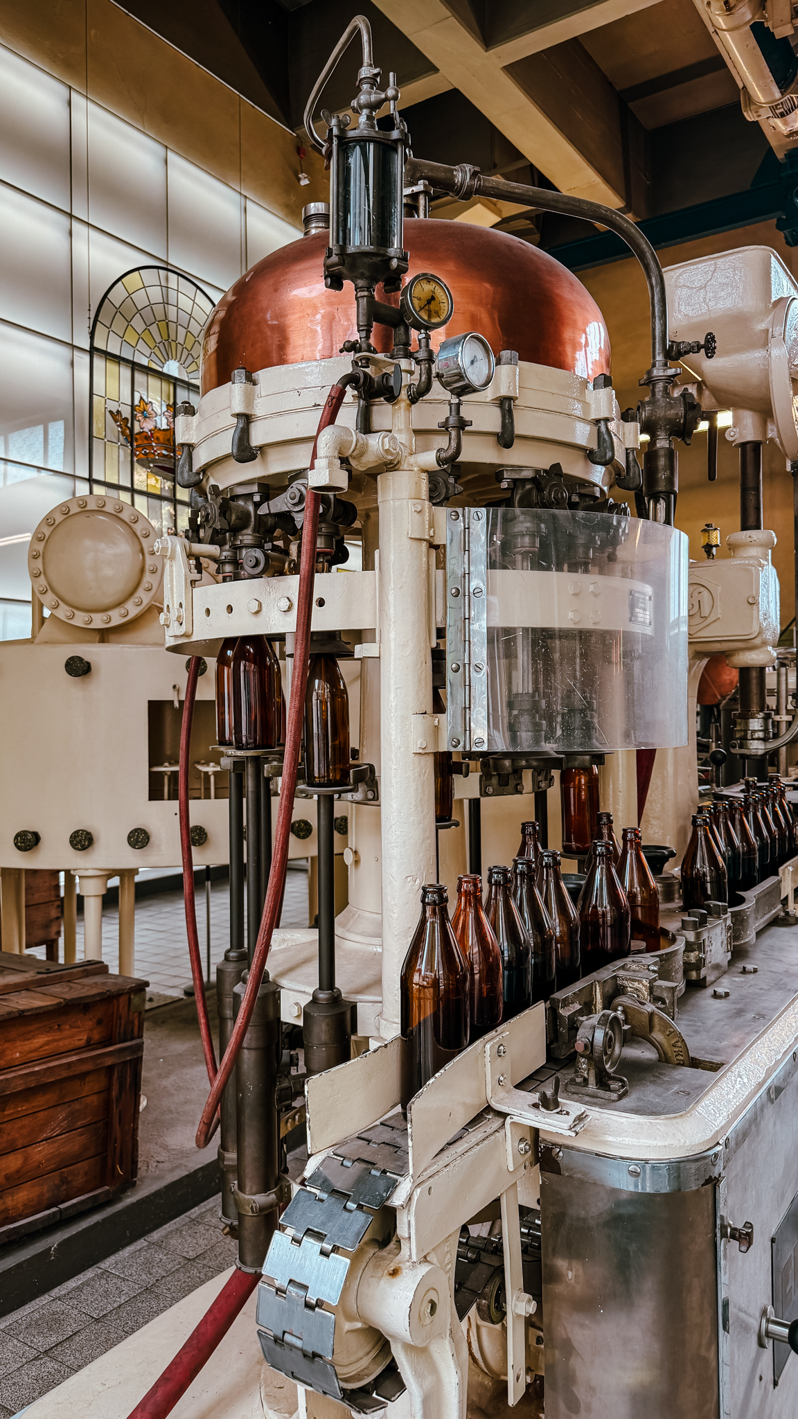 Eine alte industrielle Abfüllmaschine mit braunen Glasflaschen auf einem Förderband, einem Kupfertank darüber und mechanischen Komponenten in einer gut beleuchteten Fabrikumgebung.