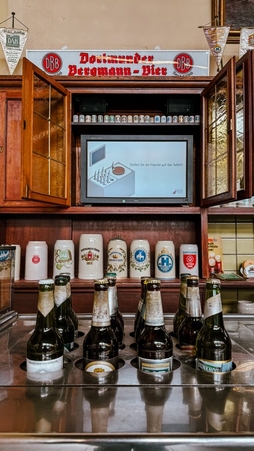 Eine Gruppe leerer Bierflaschen steht auf einem Tresen, im Hintergrund sind Bierkrüge und ein Monitor zu sehen. Auf dem Monitor ist eine Anleitung für ein Bowlingspiel in deutscher Sprache zu sehen. Ein Schild darüber wirbt für Bergmann-Bier.