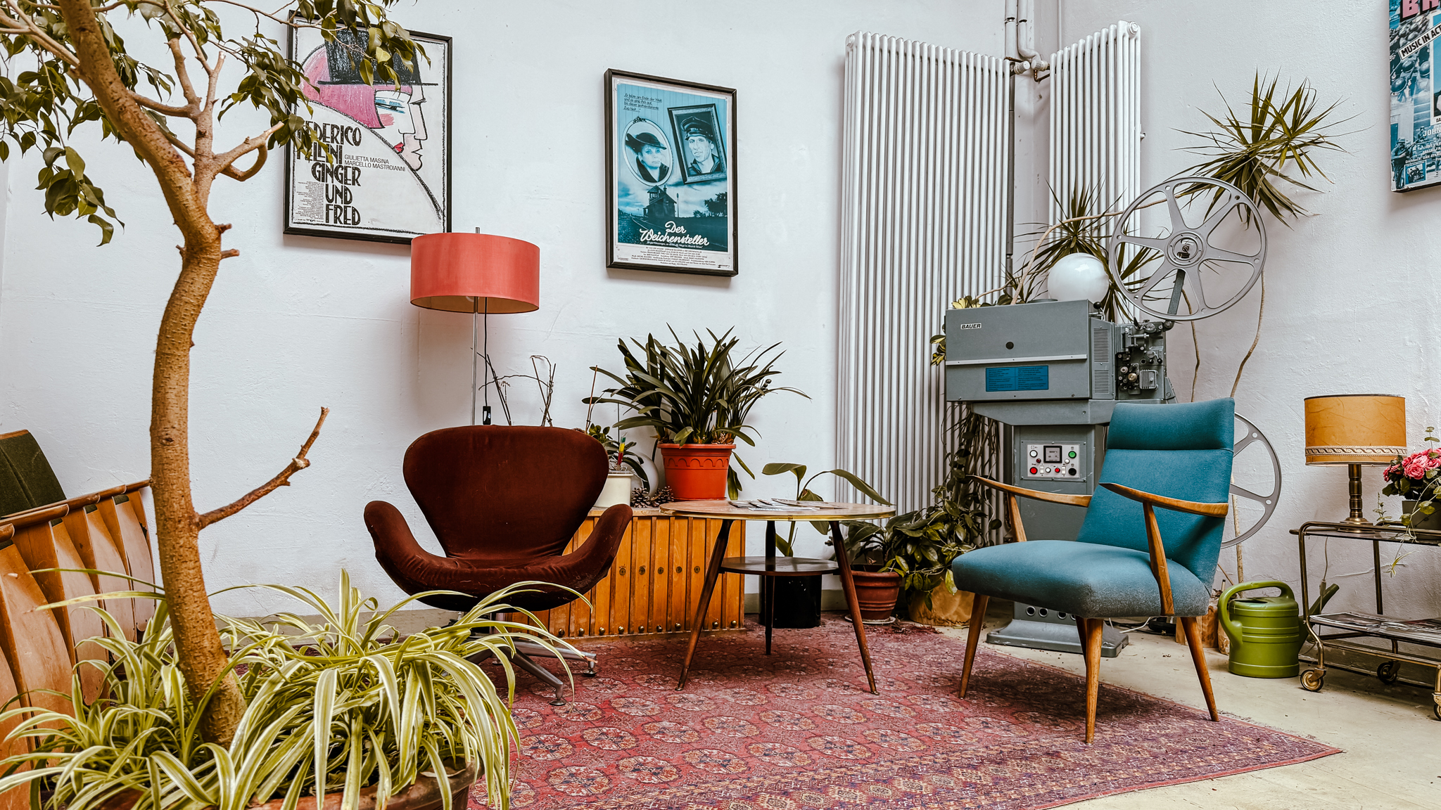 Ein gemütlicher Raum im Retrostil mit Stühlen aus der Mitte des Jahrhunderts, einem alten Filmprojektor, Pflanzen, gerahmten Postern an der Wand, einem roten Teppich und einer Stehlampe, die eine warme, eklektische Atmosphäre schaffen.