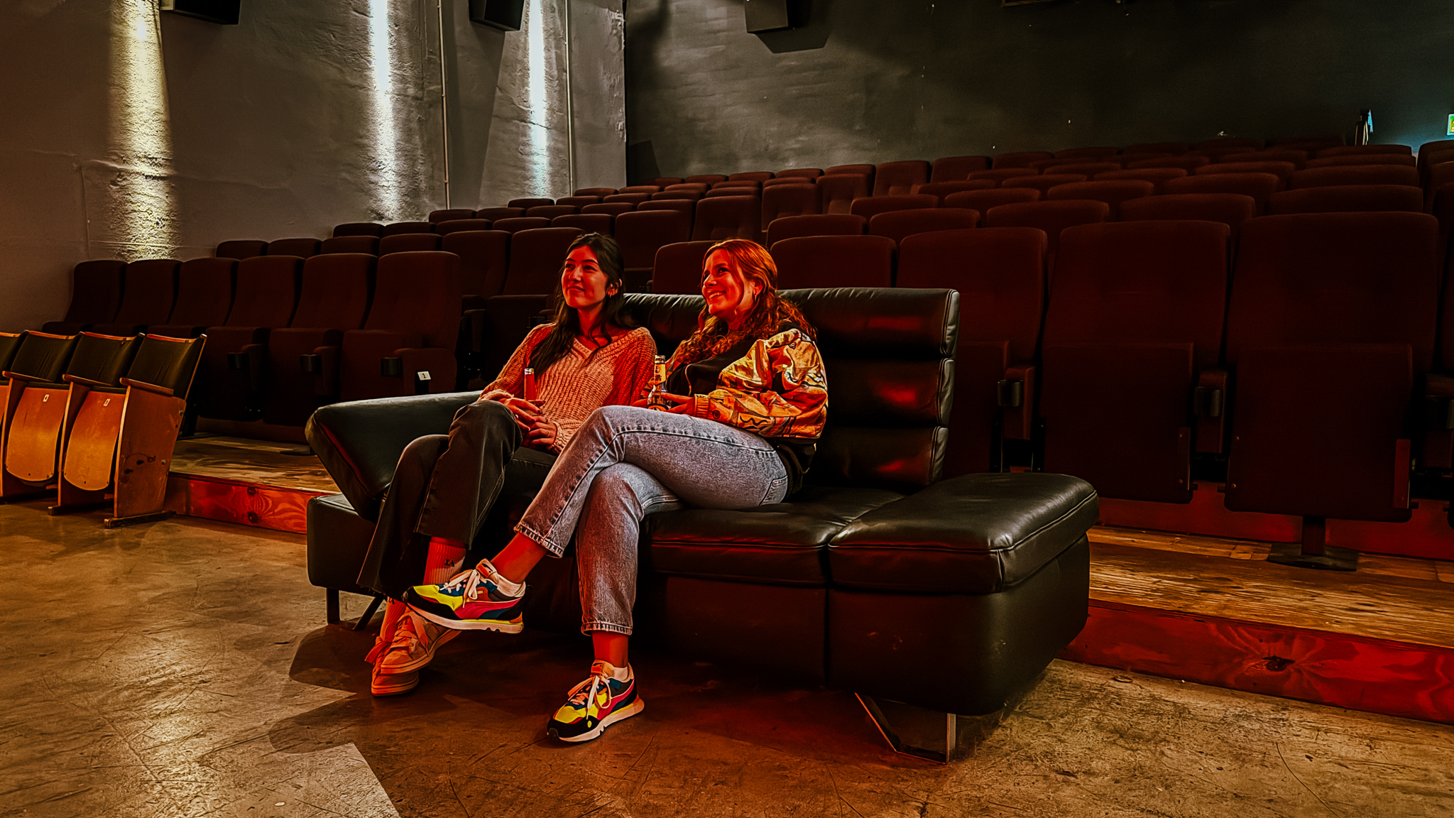Zwei Personen sitzen auf einer schwarzen Couch in einem leeren Kino und schauen auf die Leinwand. Der Raum ist schwach beleuchtet, und hinter ihnen sind Reihen von leeren roten Sitzen zu sehen.