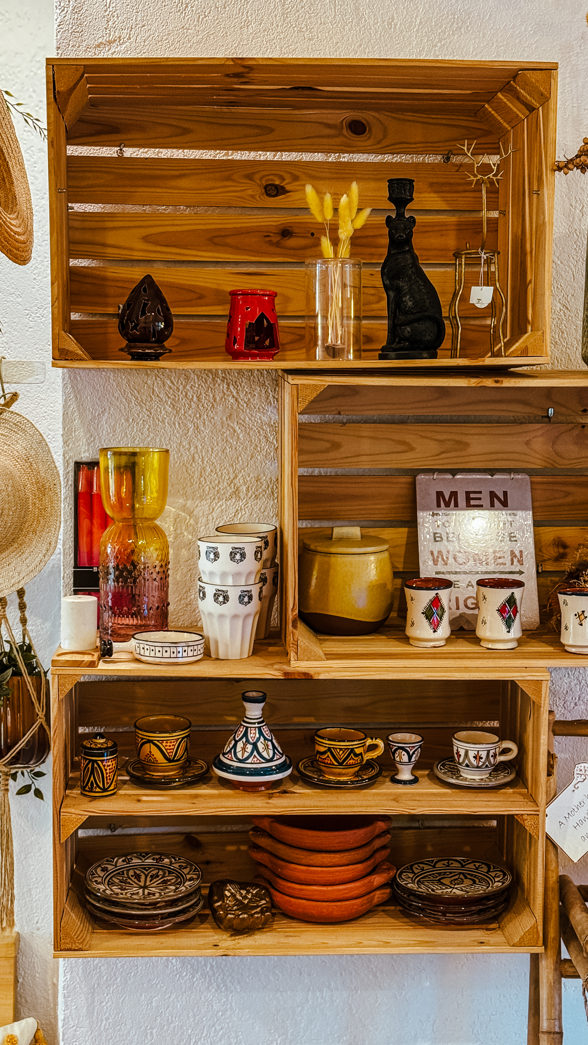 In den Holzregalen stehen bunte Gläser, Töpferwaren, Tassen, Schalen, dekorative Teller, Kerzen, ein Schild mit der Aufschrift MEN zur Linken WOMEN zur Rechten und kleine Trockenblumenarrangements vor einer weißen Wand.