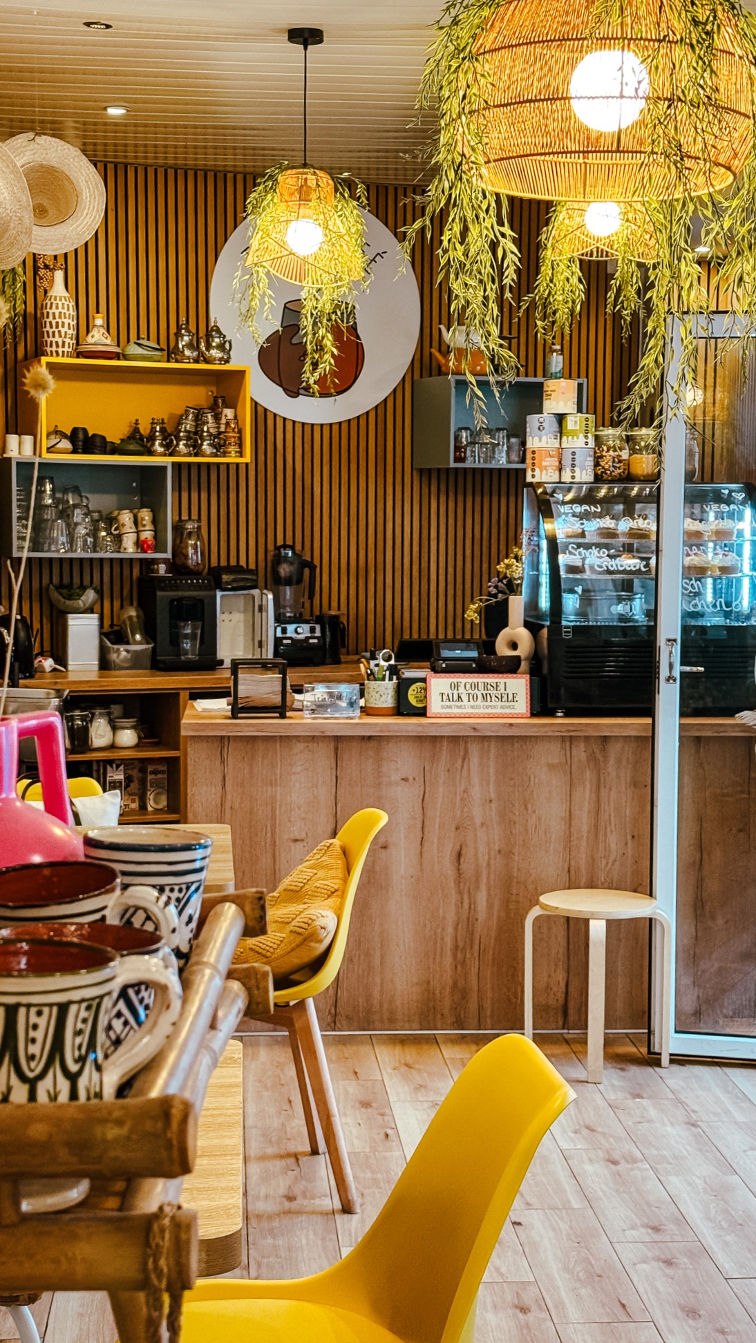 Ein gemütliches Café mit Holzwänden, hängenden Pflanzen, warmer Beleuchtung, gelben Stühlen und einer Theke mit Gebäck und Kaffeezubehör. In den Regalen stehen Tassen und Teekannen, die eine einladende und stilvolle Atmosphäre schaffen.