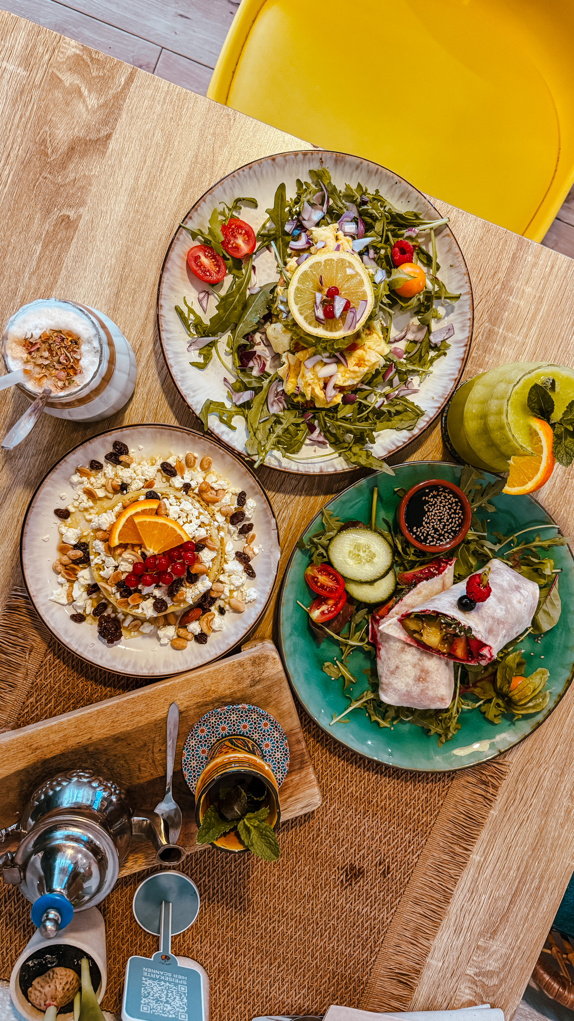 Draufsicht auf einen Tisch mit bunten, gesunden Speisen, darunter ein Wrap mit Grünzeug, ein Salat mit Zitronenscheiben, eine Schüssel Joghurt mit Früchten und Nüssen, eine Teekanne und Getränke. In der Ecke ist ein gelber Stuhl zu sehen.