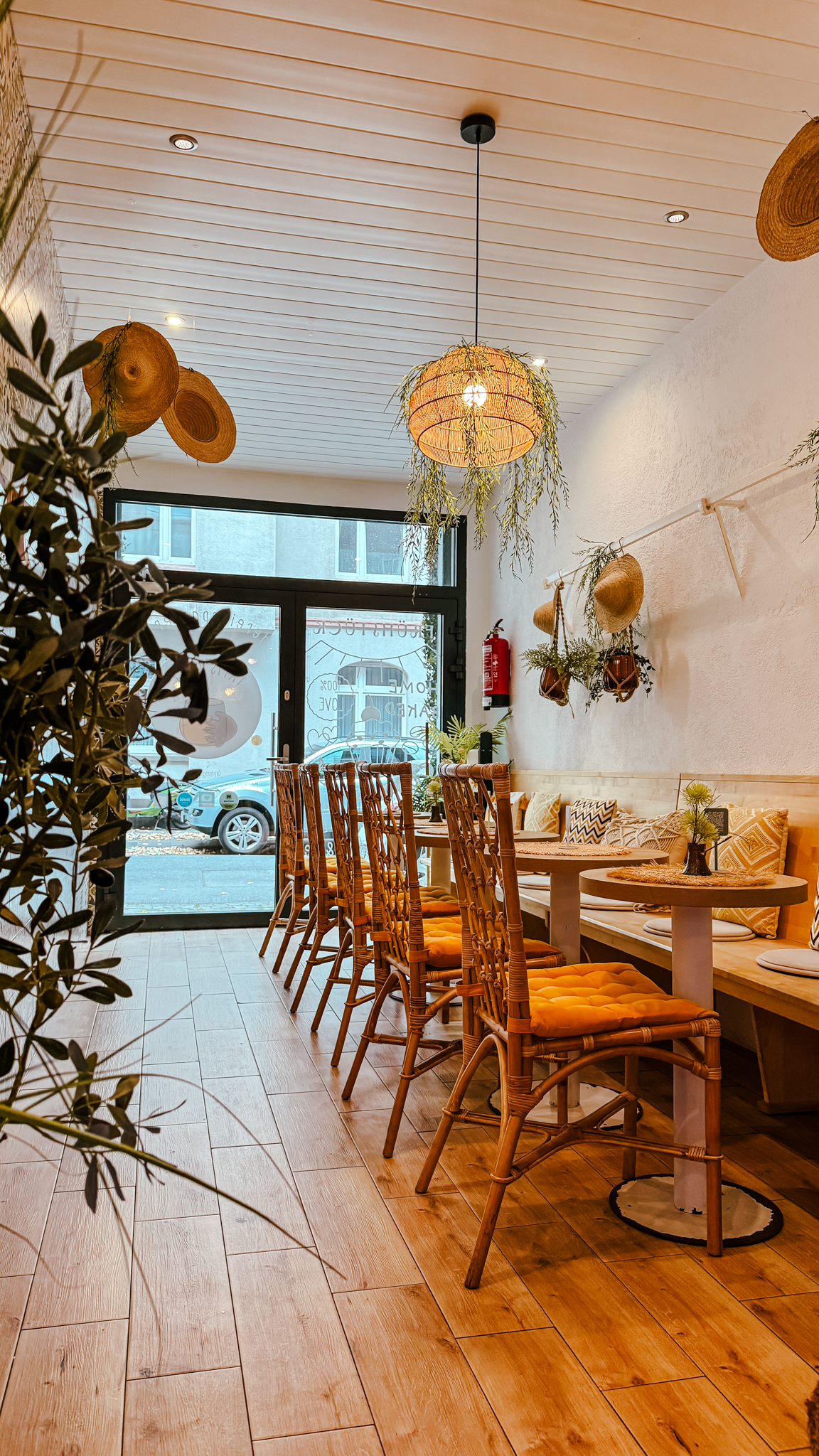 Ein gemütliches Café mit Holzböden, einem langen Tisch mit Korbstühlen, Hängelampen und rustikalem Dekor. Große Fenster lassen natürliches Licht herein, und die Bepflanzung verleiht dem einladenden Raum eine frische Note.