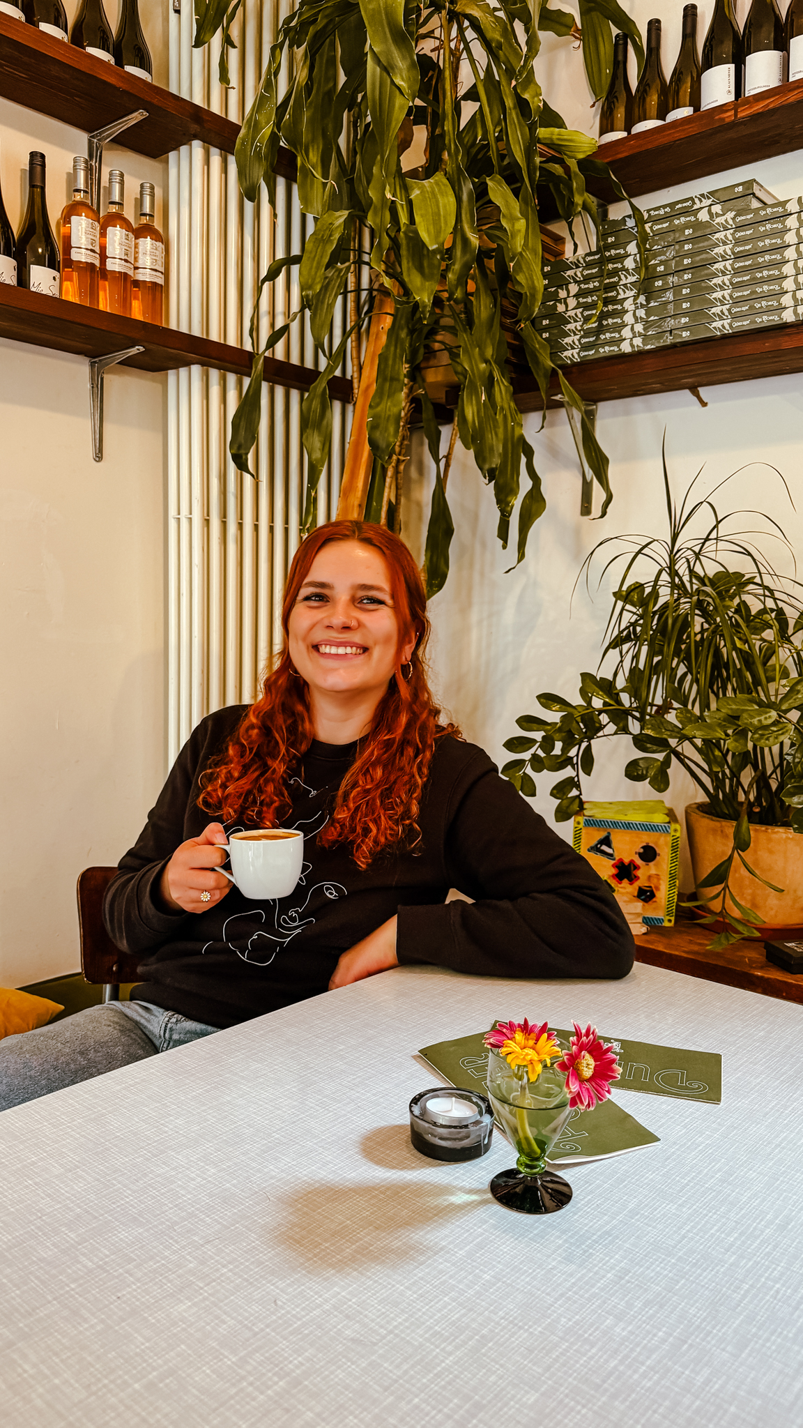 Eine lächelnde Person mit langen, gewellten roten Haaren sitzt an einem Tisch und hält einen weißen Becher. Auf dem Tisch stehen Blumen, in den Regalen Pflanzen und an der Wand hinter ihnen Flaschen in einer gemütlichen Café-Atmosphäre.