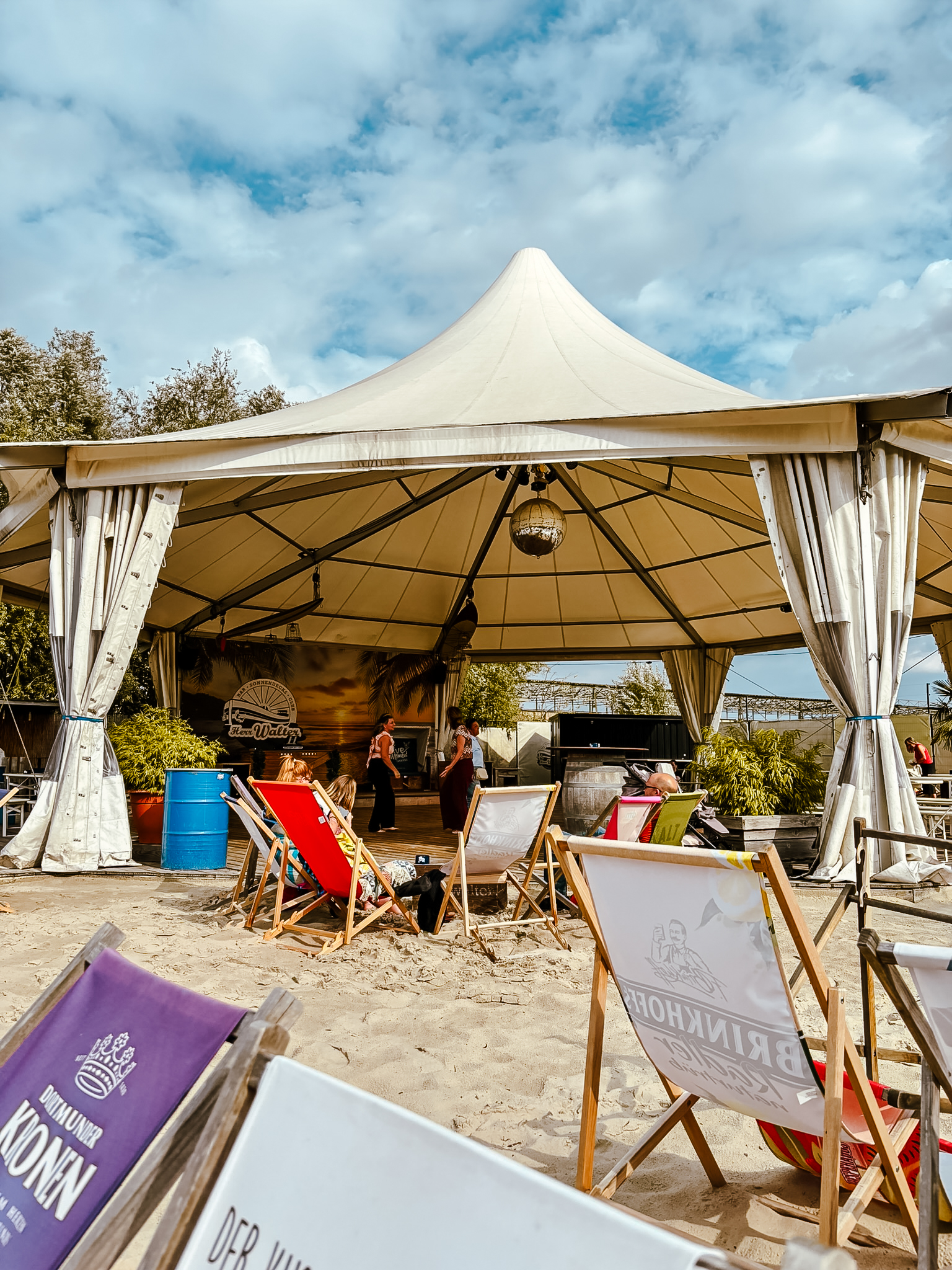 Ein großes weißes Zelt mit einer Bühne darunter ist auf einer Sandfläche aufgebaut. Im Vordergrund sind bunte Liegestühle aufgestellt, und in der Nähe der Bühne stehen ein paar Leute. Der Himmel ist teilweise bewölkt.