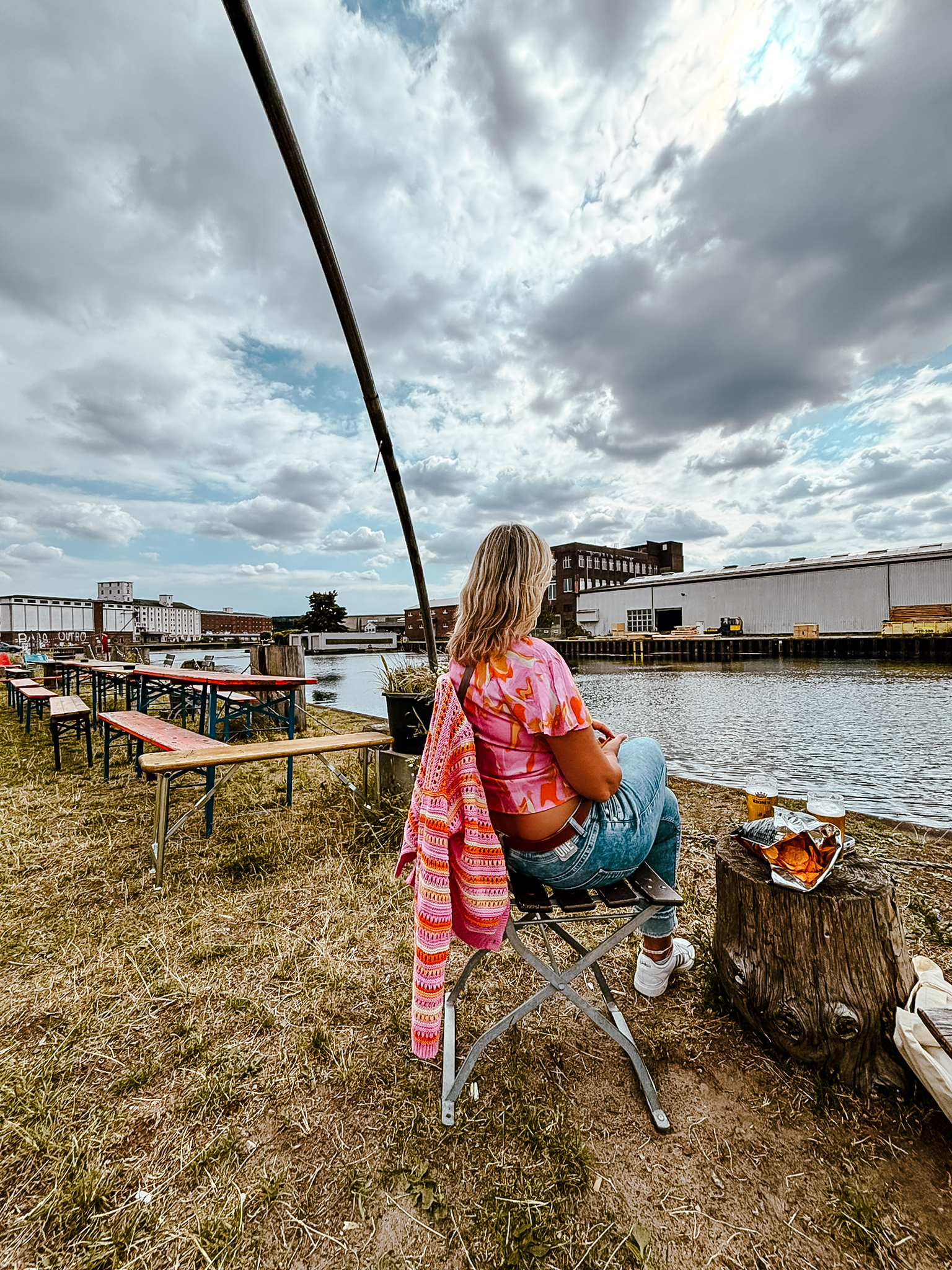 Eine Person mit gewelltem blondem Haar sitzt auf einer Bank mit Blick auf den Fluss, sie trägt ein rosa Batikhemd und Jeans. Leere Picknicktische und Bänke säumen das grasbewachsene Flussufer unter einem bewölkten Himmel. Snacks liegen in der Nähe auf einem Baumstumpf.