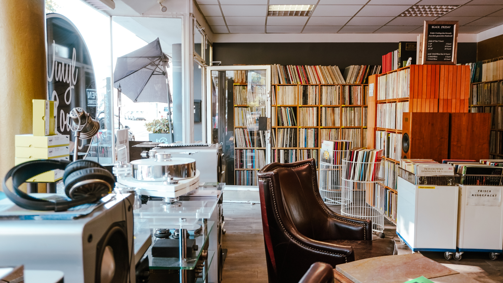 Ein gemütlicher Plattenladen mit Regalen voller Vinyl-Schallplatten, einem Ledersessel, Plattenspielern, Kopfhörern und Plattenwägen. Durch ein großes Fenster strömt Sonnenlicht herein und schafft eine warme und einladende Atmosphäre.