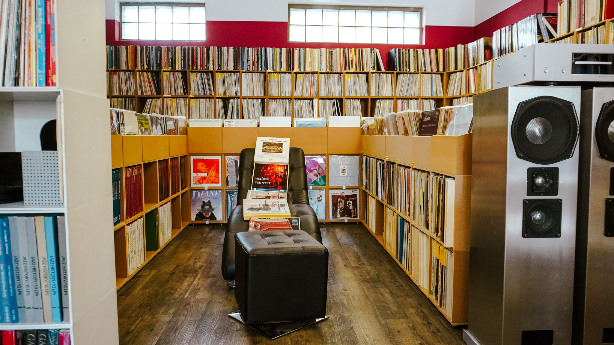 Ein gemütlicher Plattenladen mit Regalen voller Schallplatten, einem schwarzen Stuhl und einer Ottomane in der Mitte und großen Lautsprechern auf der rechten Seite. Der Raum hat Holzböden und das Sonnenlicht fällt durch die hohen Fenster.