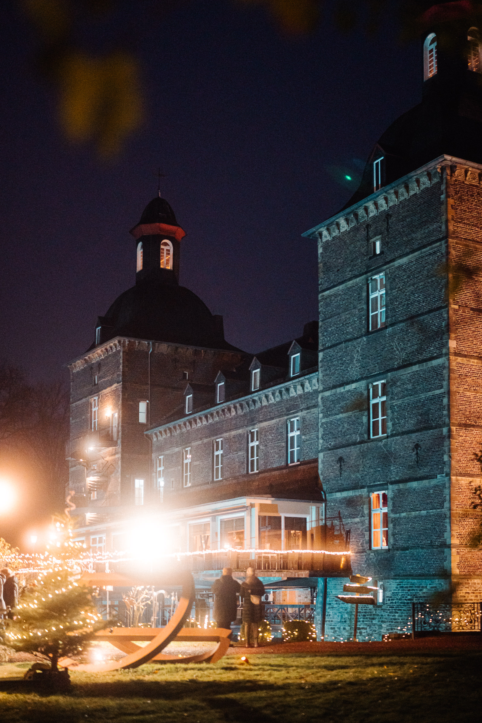 Eine nächtliche Szene eines großen, historischen Backsteingebäudes mit Türmen, warm beleuchteten Fenstern und einem festlich geschmückten Außenbereich mit Lichterketten und einem kleinen beleuchteten Baum. Menschen stehen in der Nähe des Eingangs.