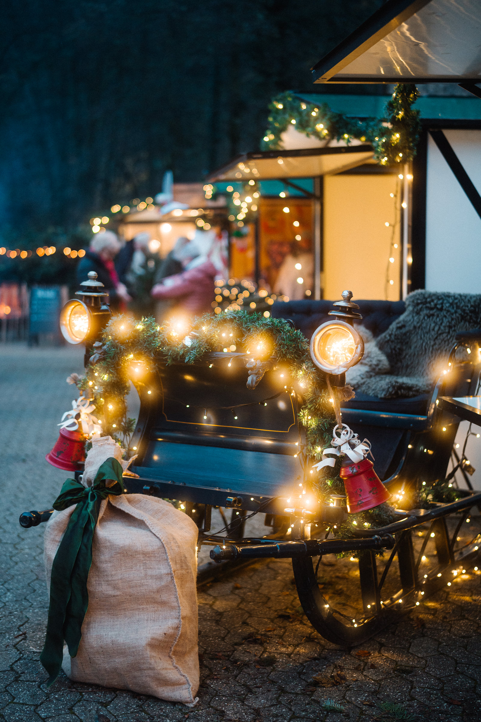 Ein festlich geschmückter Schlitten mit Lichterketten, Girlanden und roten Glocken steht neben einem großen Sack aus Sackleinen, der mit grünem Band verschnürt ist, auf einem Weihnachtsmarkt mit beleuchteten Verkaufsständen im Hintergrund.