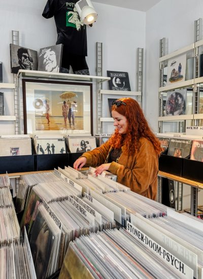 Eine Frau mit langen roten Haaren stöbert in einem Plattenladen in Vinylplatten, umgeben von Regalen mit Alben und musikbezogener Dekoration.