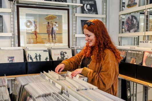 Eine Frau mit langen roten Haaren stöbert in einem Plattenladen in Vinylplatten, umgeben von Regalen mit Alben und musikbezogener Dekoration.