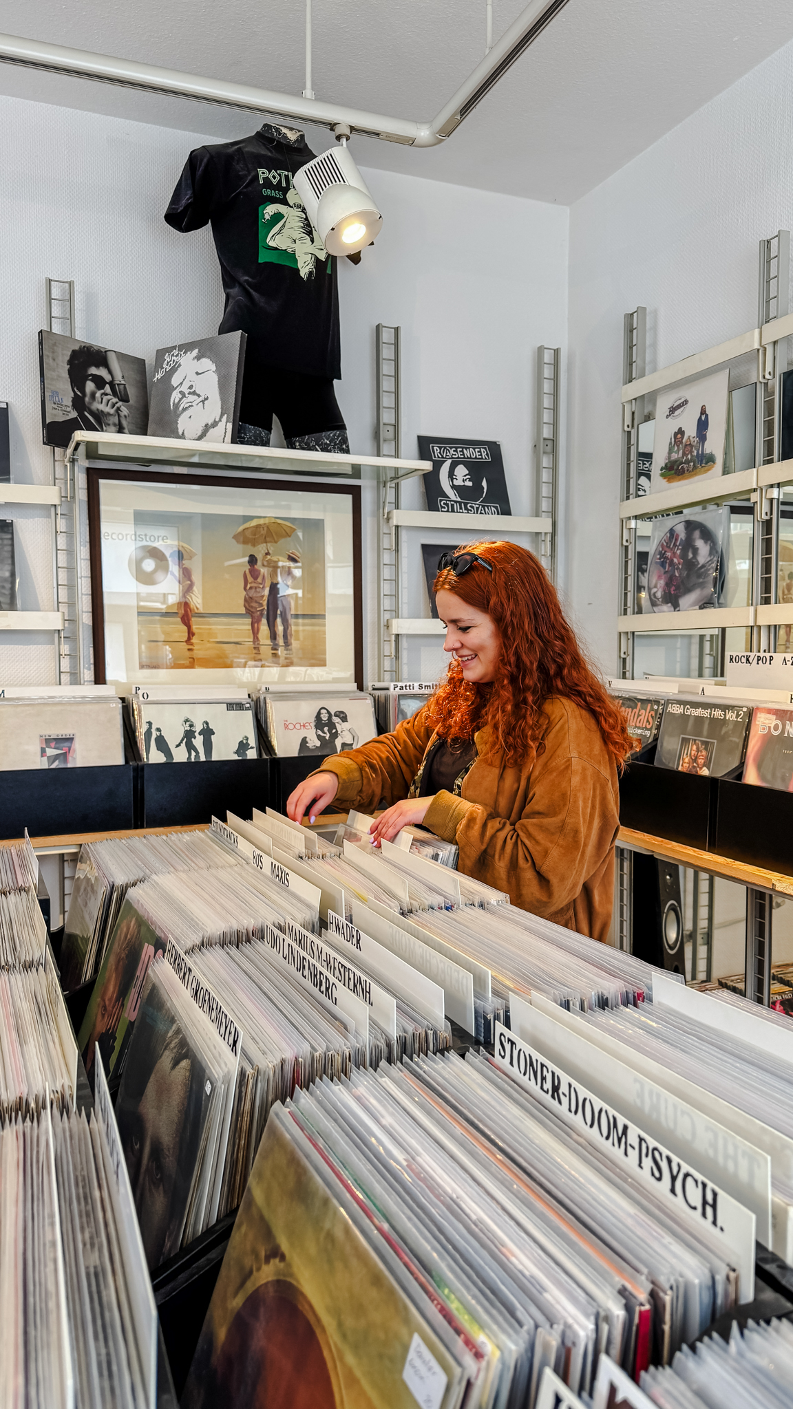 Eine Frau mit langen roten Haaren stöbert in einem Plattenladen in Vinylplatten, umgeben von Regalen mit Alben und musikbezogener Dekoration.