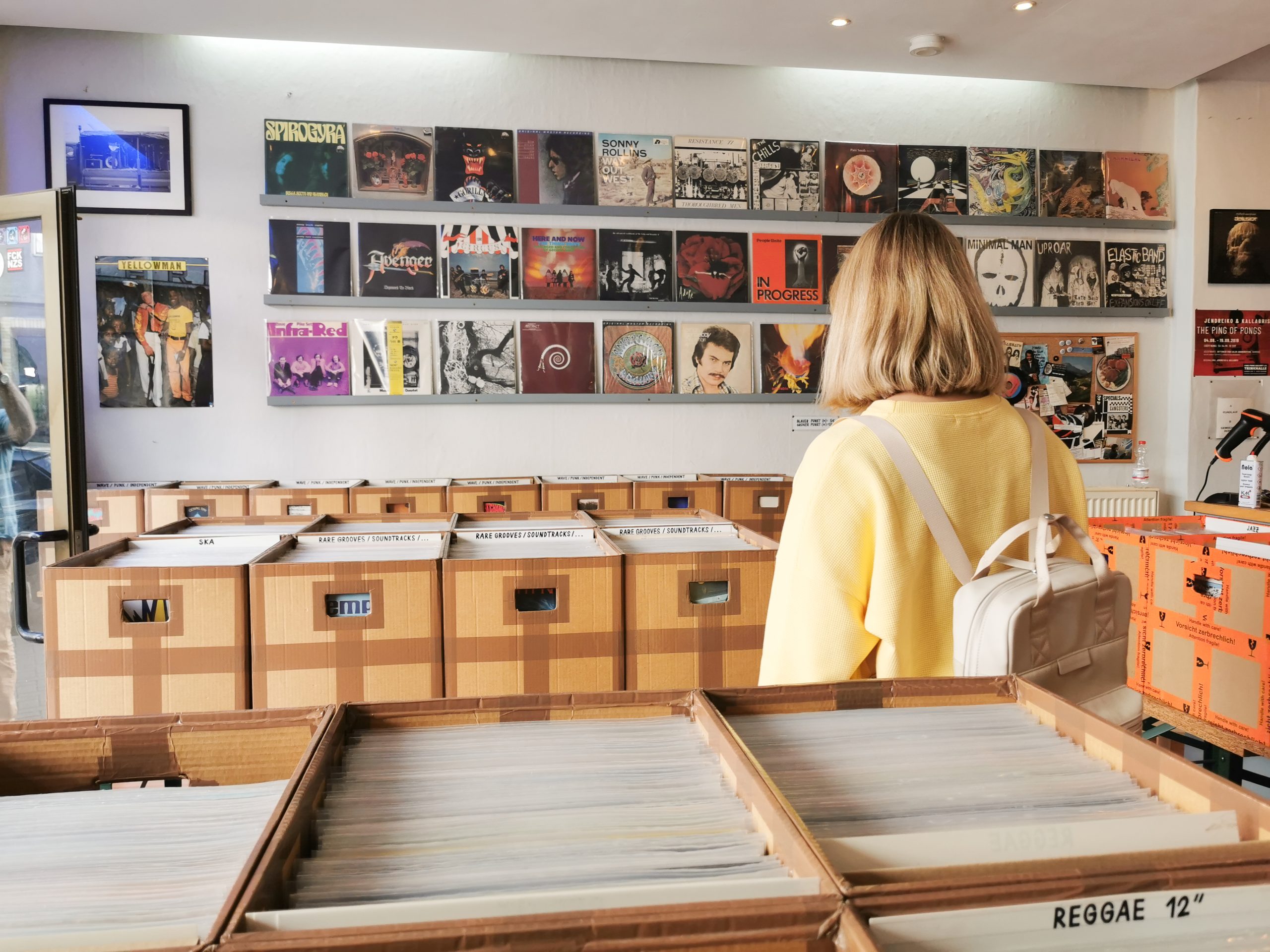 Eine Person mit blondem Haar, die ein gelbes Oberteil und einen weißen Rucksack trägt, stöbert in einem Plattenladen in Schallplatten. An der Wand hängen Plattencover und auf den Tischen stehen Kartons mit Schallplatten.