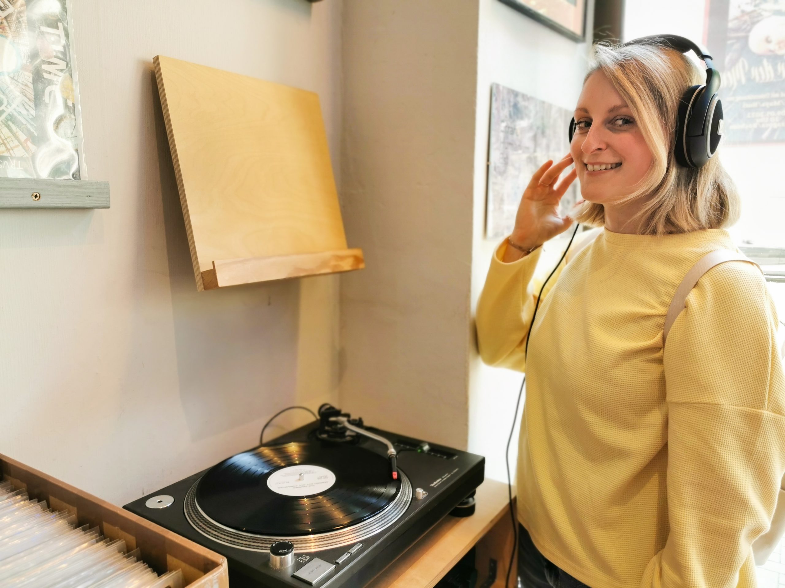 Eine lächelnde Frau mit Kopfhörern steht neben einem Schallplattenspieler in einem gemütlichen Raum und scheint Musik zu hören. Sie ist mit einem gelben Pullover bekleidet und blickt in die Kamera.