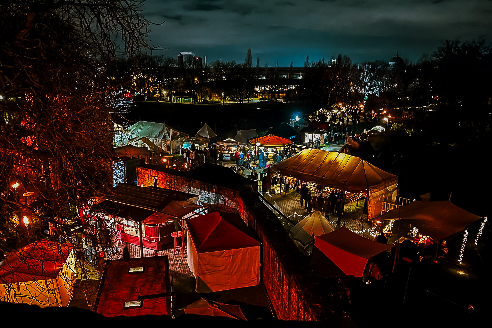 Ein nächtlicher Markt im Freien mit bunt beleuchteten Zelten und Lichterketten. Menschen gehen zwischen den Ständen umher, und im dunklen Hintergrund sind Bäume und Gebäude unter einem bewölkten Himmel zu sehen.