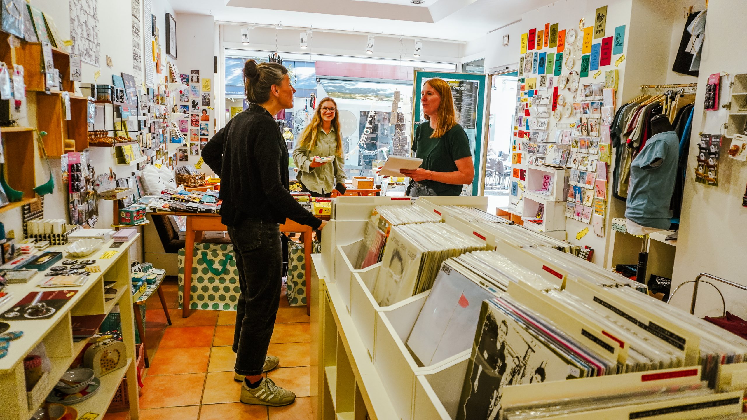 Drei Frauen plaudern und lächeln in einem farbenfrohen, gut beleuchteten Laden, der mit Schallplatten, Büchern und verschiedenen kleinen Geschenkartikeln auf Regalen und Tischen gefüllt ist.