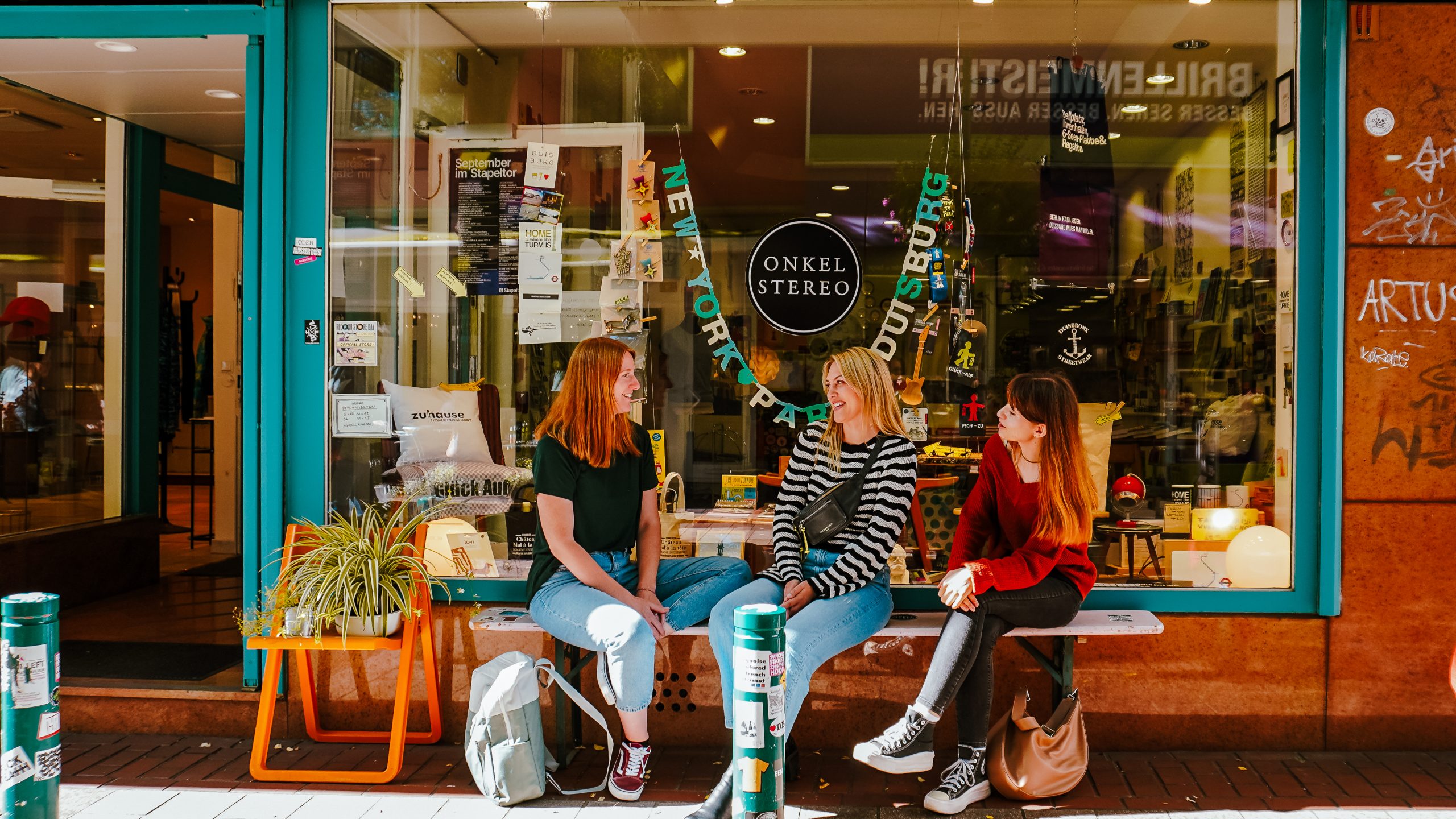 Drei junge Frauen sitzen und unterhalten sich vor einem Geschäft namens "Onkel Stereo", umgeben von Taschen und Grünzeug. Das Schaufenster ist mit Postern und buntem Dekor bedeckt. Die Straße ist in Orange- und Grüntönen gehalten.