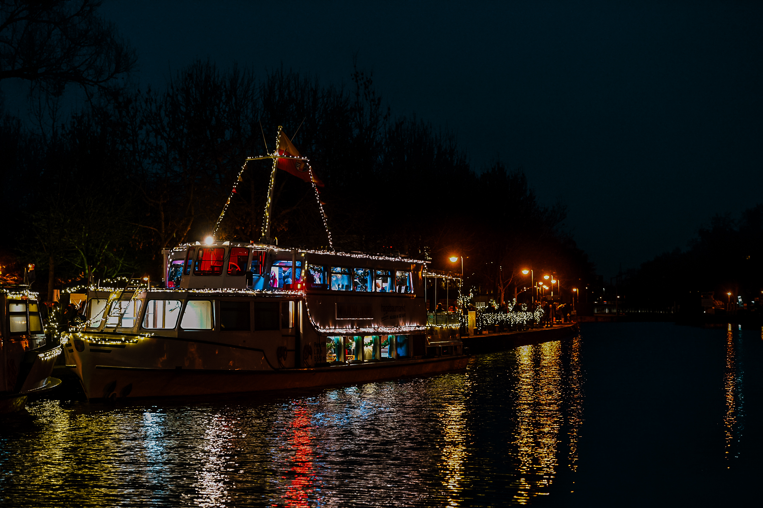Ein hell erleuchtetes, mit Lichterketten geschmücktes Boot liegt nachts an einem ruhigen Fluss und reflektiert bunte Lichter auf dem Wasser, während am Ufer Bäume und Straßenlampen zu sehen sind.